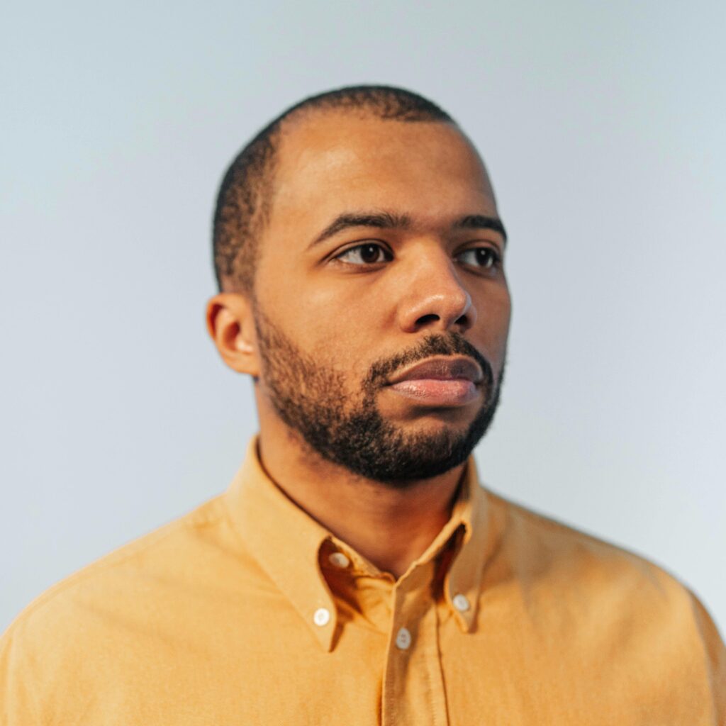 Close-up portrait of a bearded young adult man wearing a mustard shirt, set against a soft background.